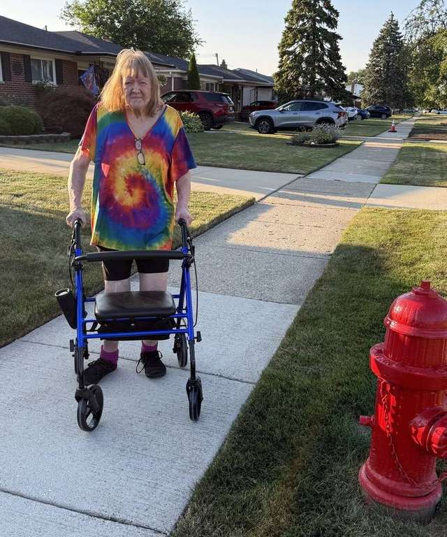 Karen smiling on a neighborhood sidewalk in a rainbow tie-dye shirt, standing tall with her blue rollator on her first walk