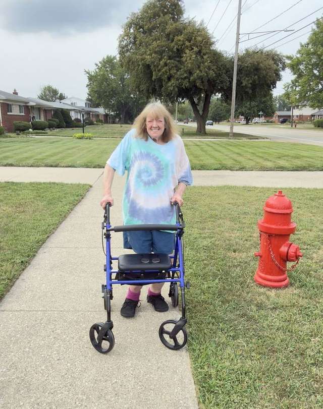 Karen out on the same sidewalk three weeks later in a blue tie-dye shirt, walking farther with her rollator and grinning